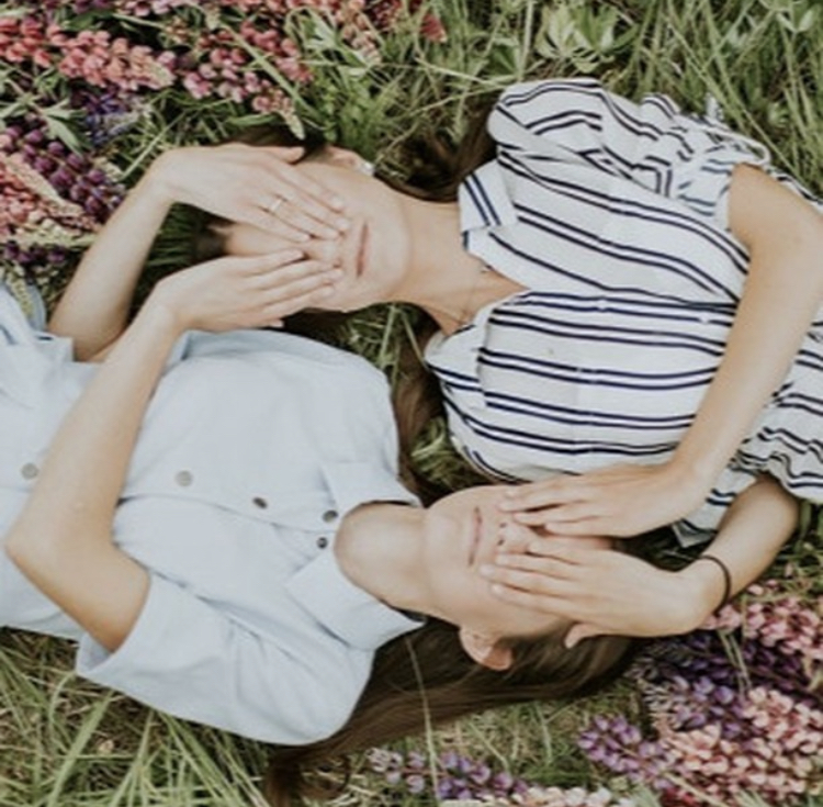 Two young women lying on their backs in the grass, their hands covering each other's eyes