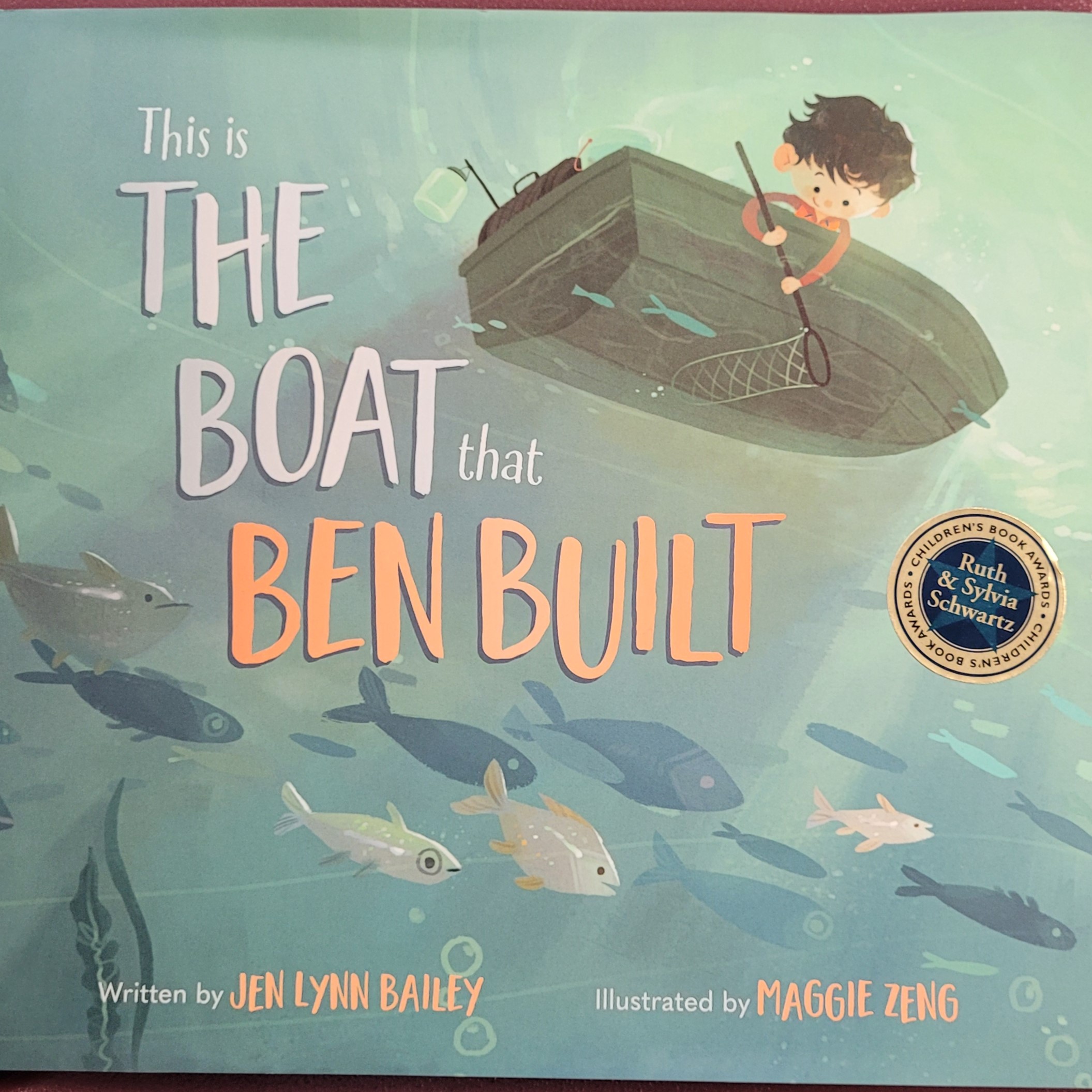 A cover of a picture book, showing a boy in a boat with a school of fish underneath him. The cover has a sticker to indicate that it won the Ruth & Sylvia Schwartz Children's Book Award.