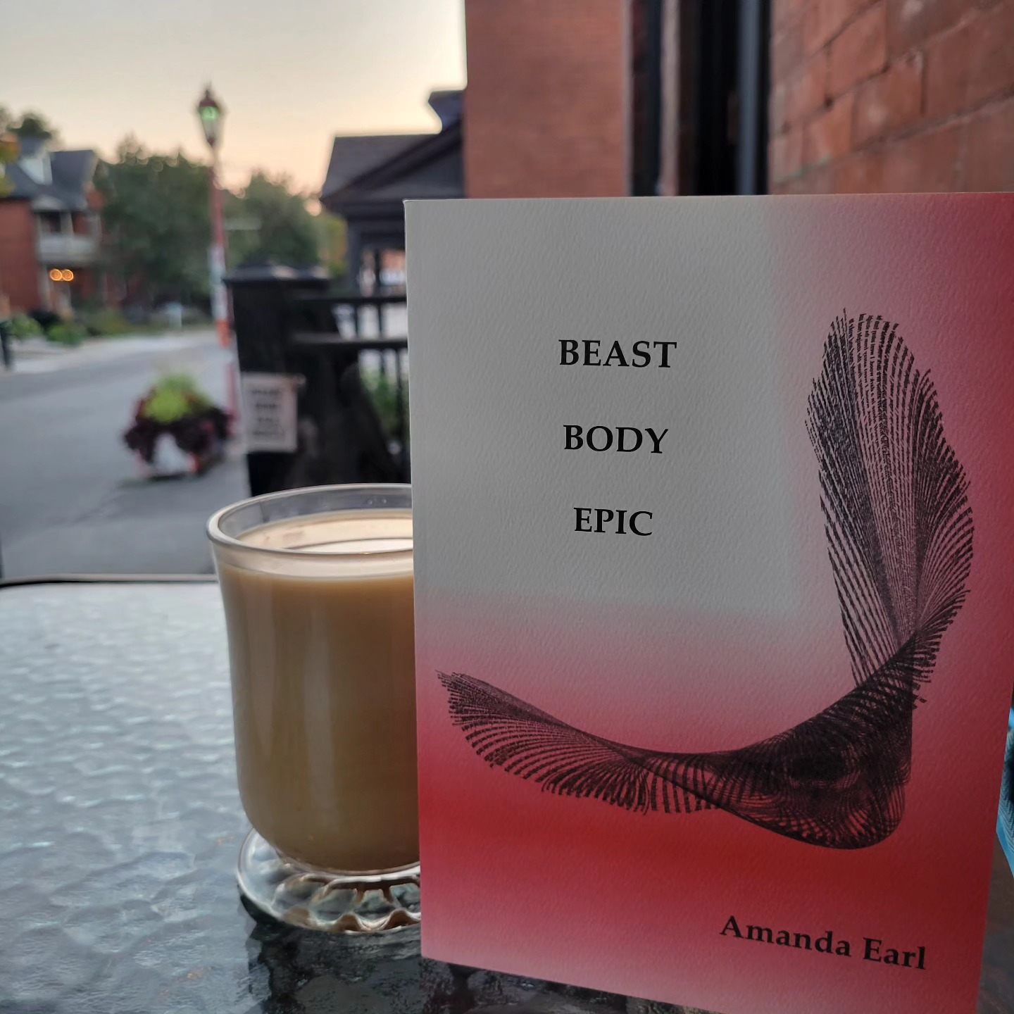 red and white book with text: Beast Body Epic / Amanda Earl and visual poem beside a cup of coffee outside on a patio, blurred background