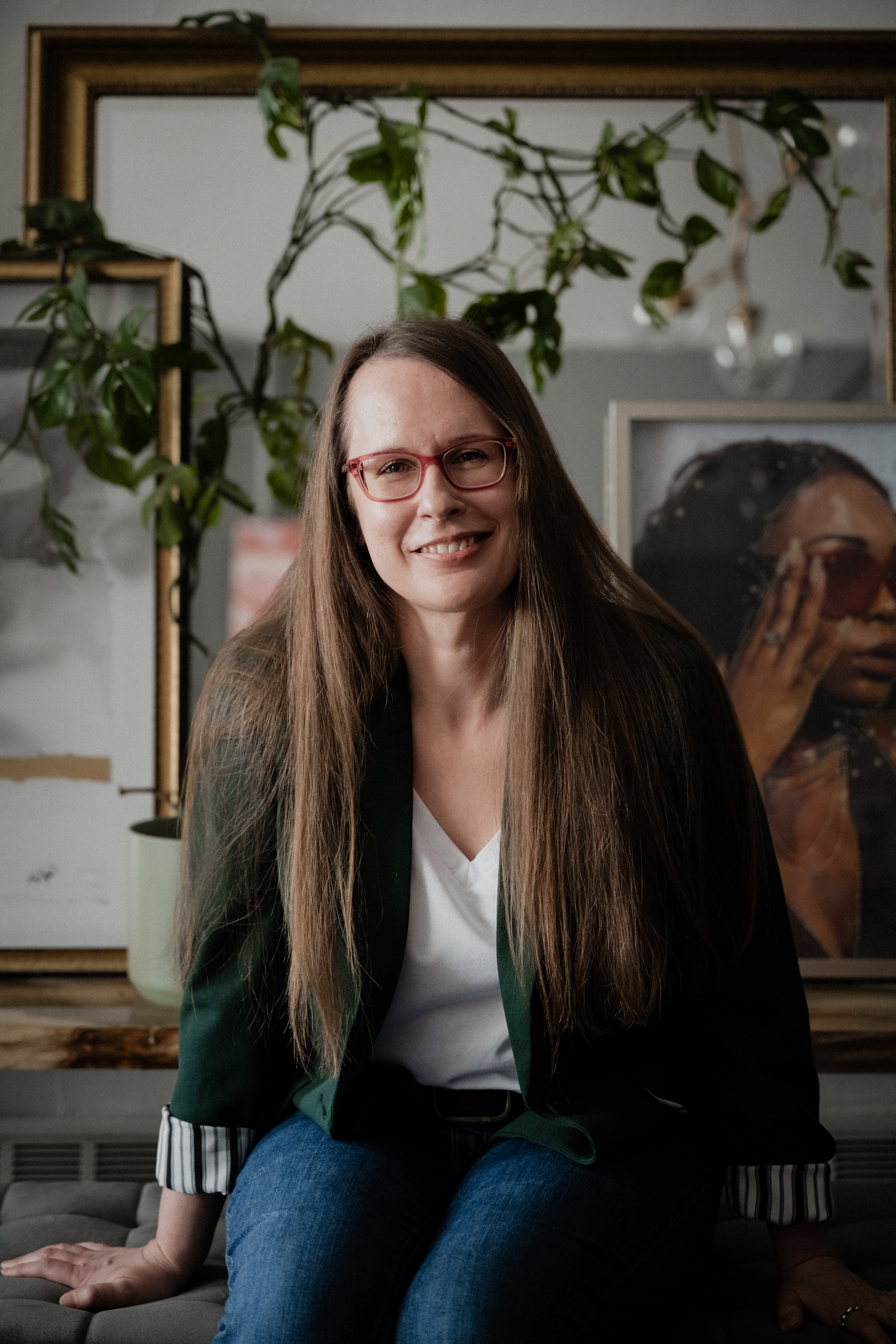 A woman seated and smiling. She has long brown hair, pink glasses and wears a green blazer and white t-shirt.