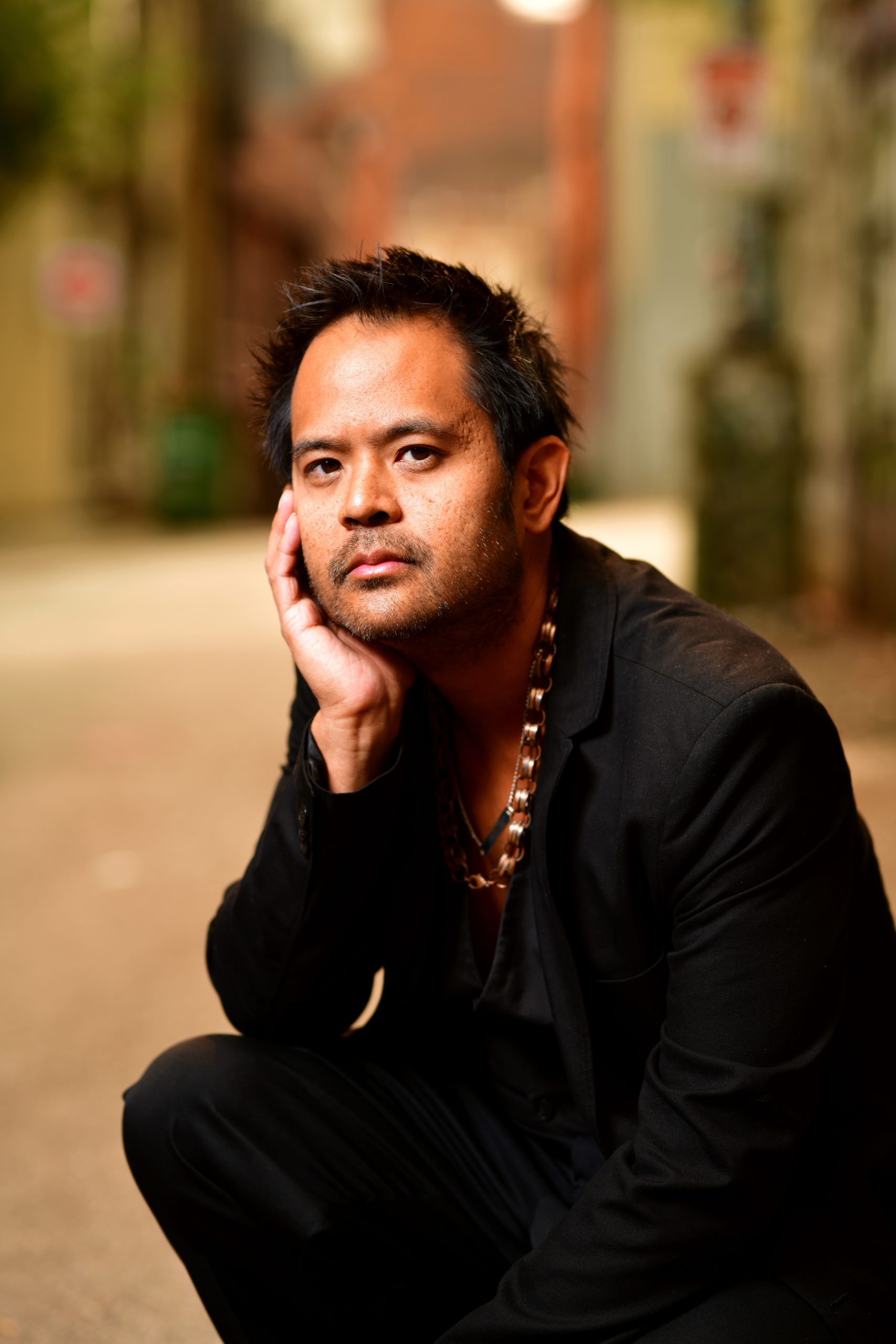 Filipino man in a black jacket and neck chains, looking at the camera, kneeling, right hand on knee and holding chin, in a Vancouver alley. 